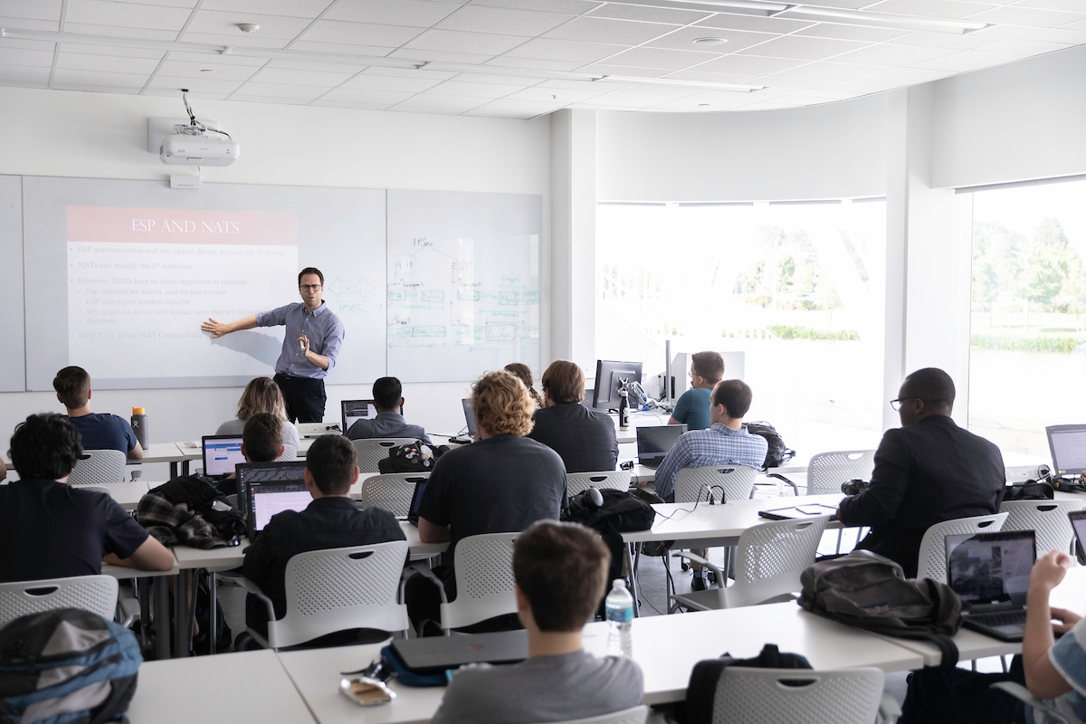 Teacher standing at white board with students sitting at tables.