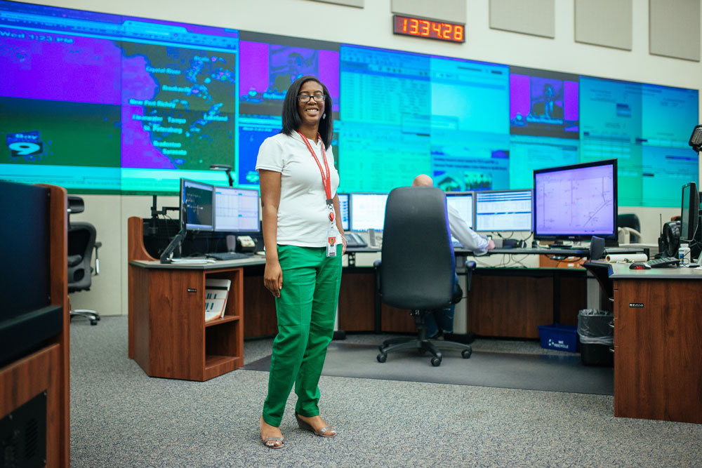 Female student in green pants stands in front of screens and clock 