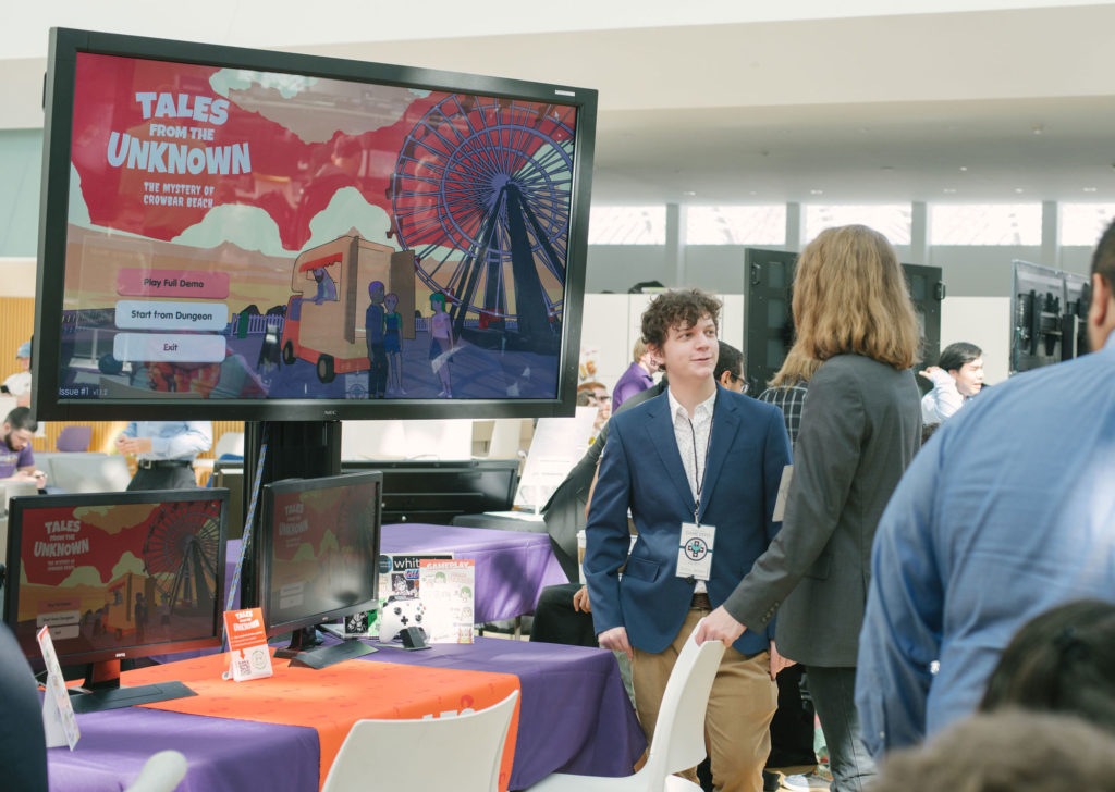 Male student in front of monitor screen explaining his game.