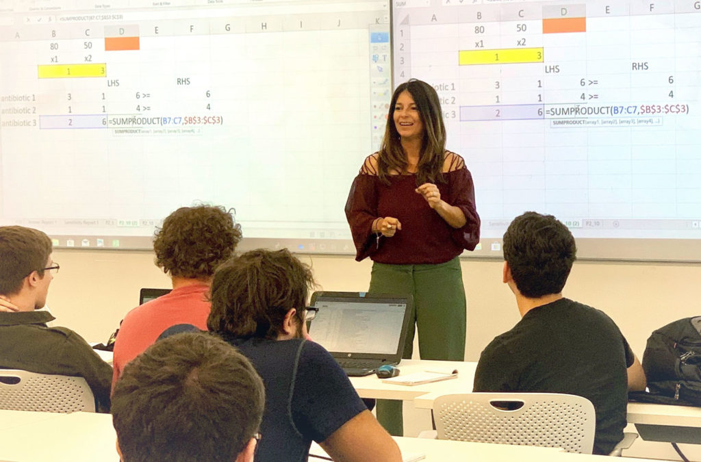 Light skinned female professor teaching in front of classroom with students.