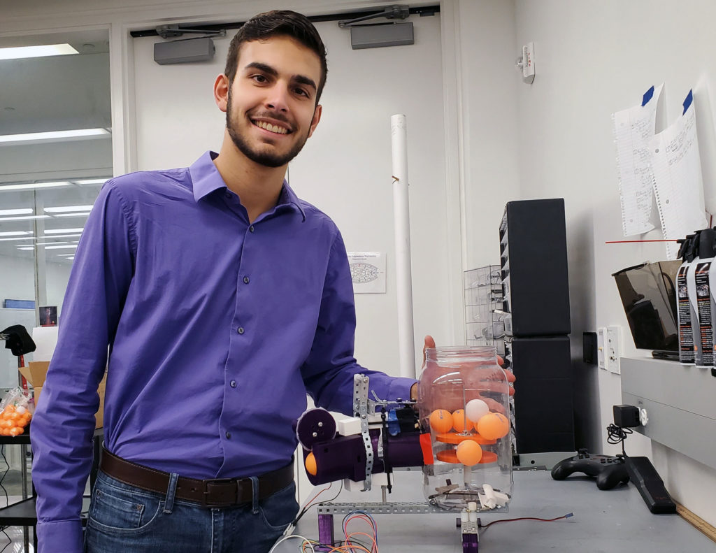 Light skinned male student standing next to ping pong machine.