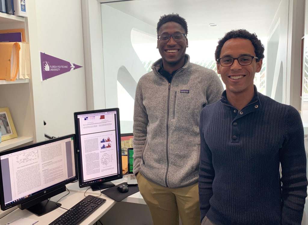 Two dark skinned males standing in front of a computer.