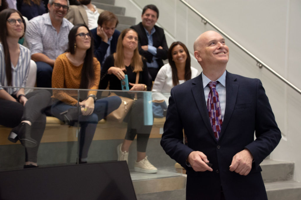 Light skinned male in a suit smiling infront of a crowd sitting.