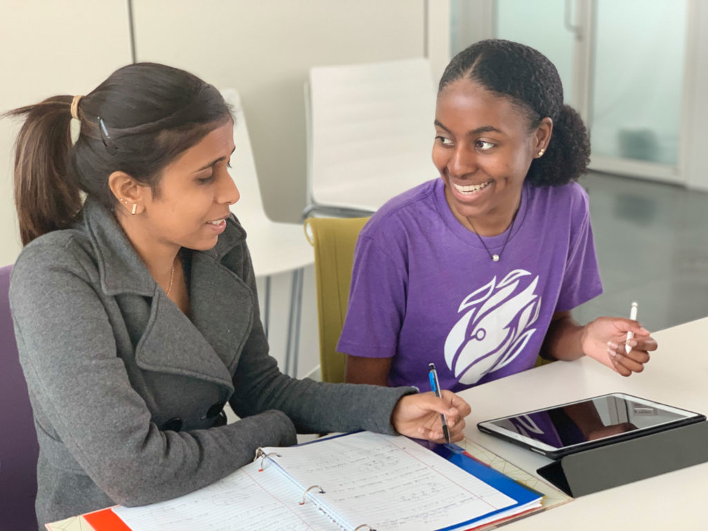 Two young women with darker skin sitting at table smiling at eachother.