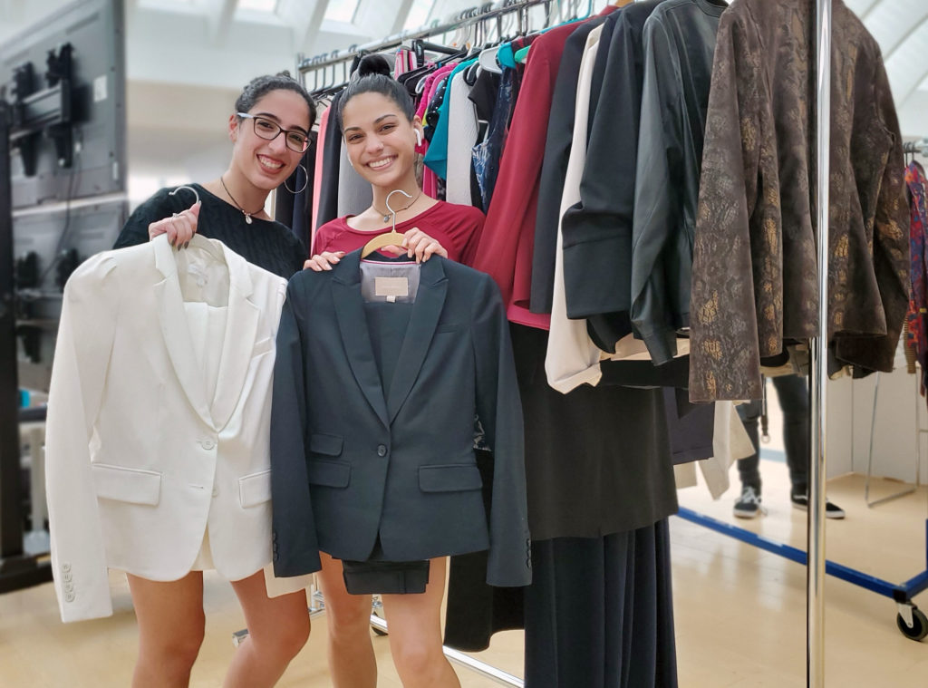 Two light skinned females holding up suits on hangers.