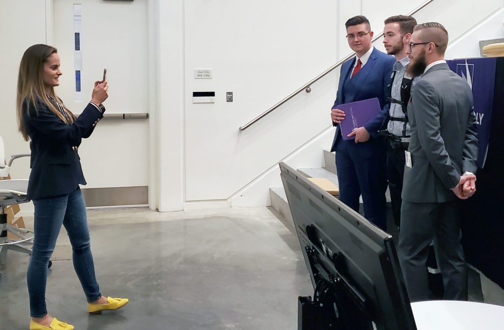 Women taking a video of three men in front of stairs.